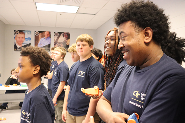 Students in Georgia Tech Advanced Manufacturing Pathways T-shirts stand in a classroom, some holding small orange objects, while others watch an activity at the front.