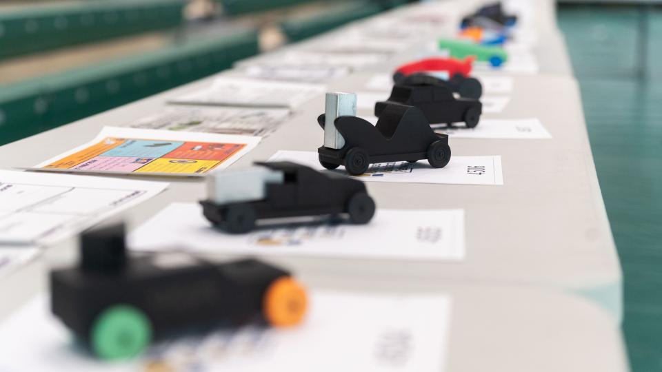 A row of small, student‑designed model race cars displayed on a table, each placed on top of design sheets in a gym setting.